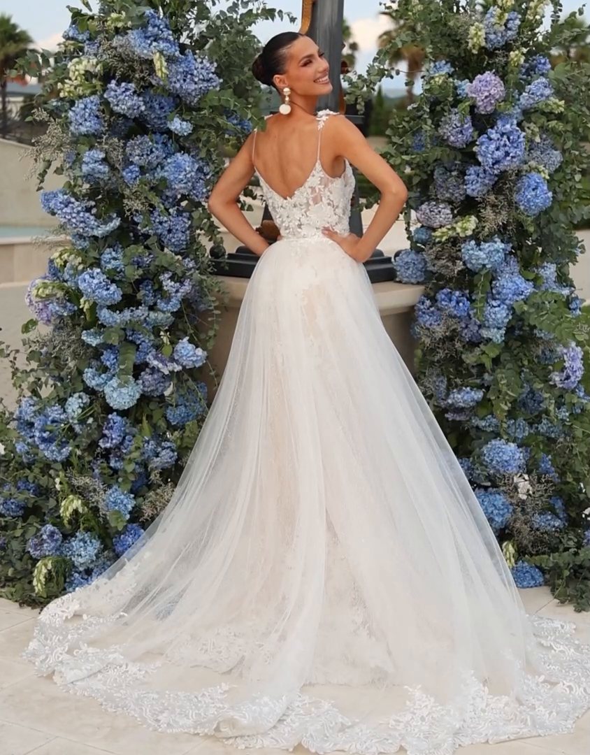 Bride in an ivory lace A-line wedding gown with a sheer embroidered back, glancing over her shoulder beside an arch of blue hydrangeas